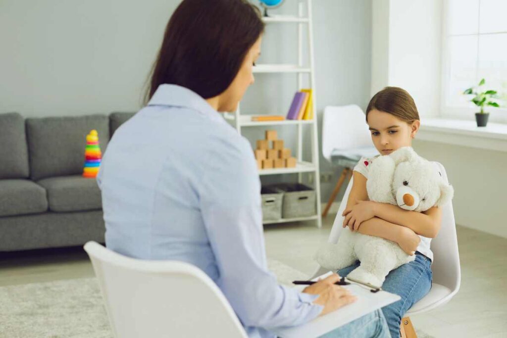 Young girl clutches a white teddy bear while sitting across from a therapist taking notes during a play-based mental health assessment in a child-focused therapy room.