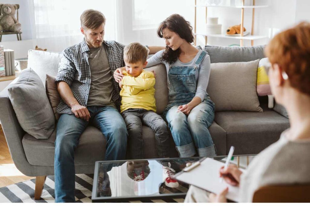 A young boy sitting between his concerned parents on a couch, arms crossed and looking down, while a therapist takes notes in the foreground during a child mental health evaluation session.