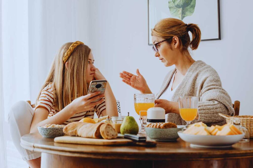 A mother and teenage daughter talk seriously at the breakfast table, showing a moment of emotional connection and open communication about feelings.