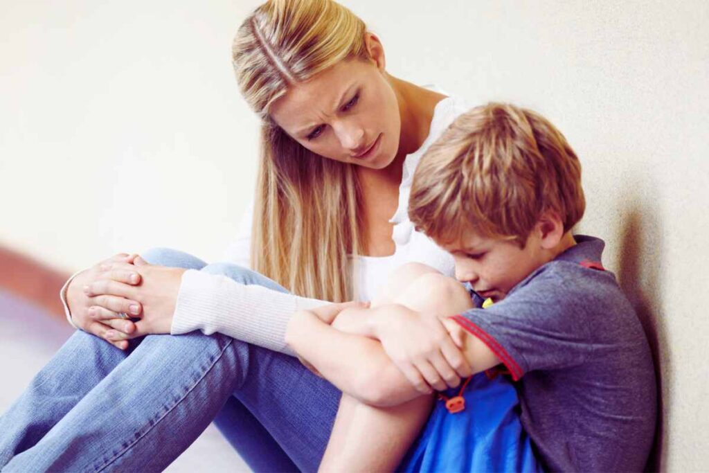 A caring mother sits beside her sad son, gently comforting him as he hides his face—capturing emotional support and safety during difficult feelings.