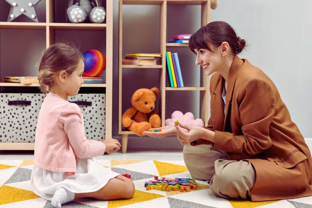 A therapist and young girl play with colorful blocks in a therapy room, illustrating the use of play to help children identify and express emotions safely.