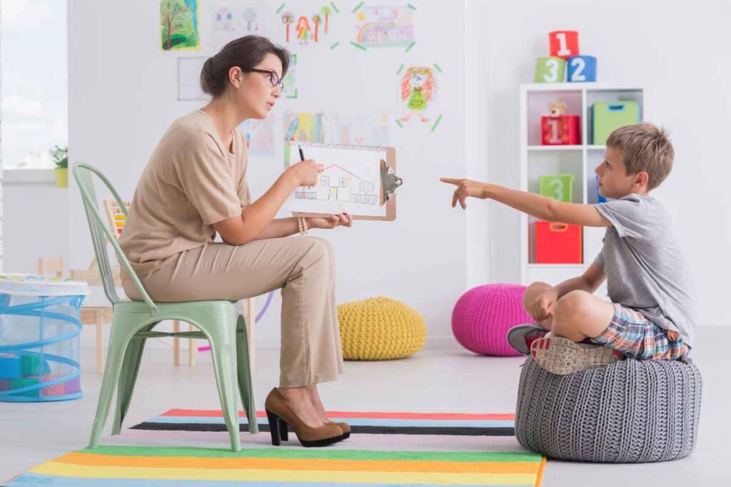 A child points at a drawing held by a therapist during a play-based mental health assessment, with colourful art and toys in the background of a child-friendly evaluation room.