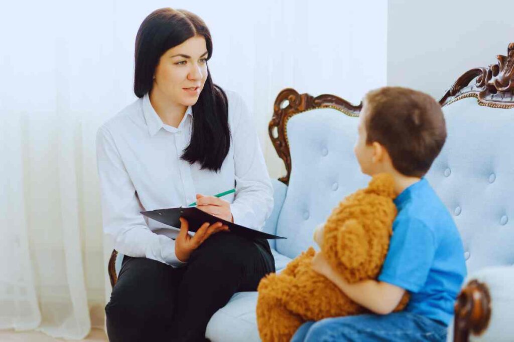 Female therapist takes notes while observing a young boy holding a teddy bear during a play-based mental health evaluation in a calm, child-friendly setting.