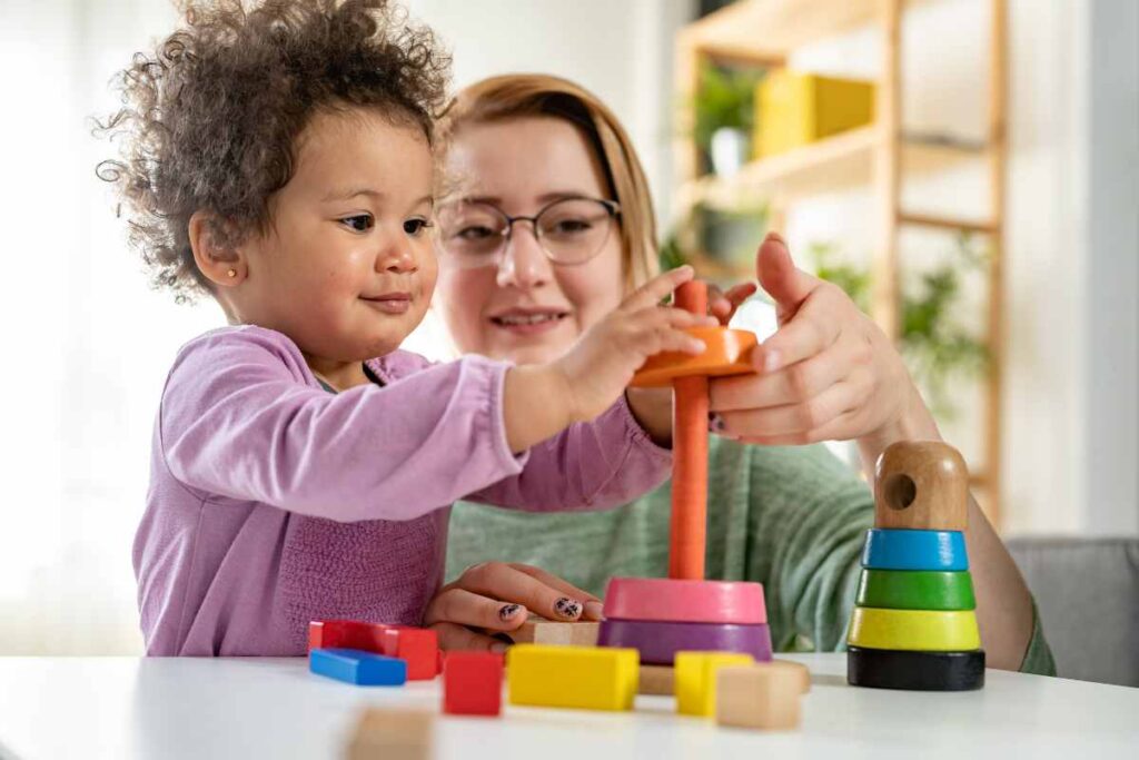 A therapist and a young child engaging in a play-based activity with stacking rings and colorful blocks, illustrating how play is used to evaluate developmental and mental health in children during a therapy session.